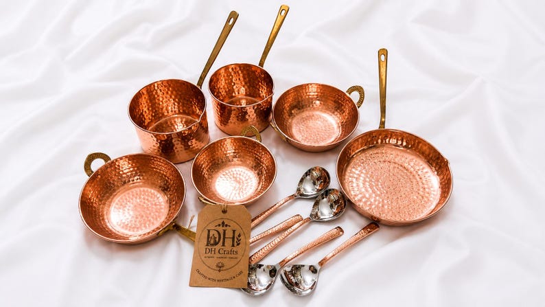 May include: A collection of hammered copper cookware including two saucepans with long handles, two bowls with side handles, one frying pan with a long handle, and four spoons. A brown paper tag with the text DH Crafts is attached to the set.