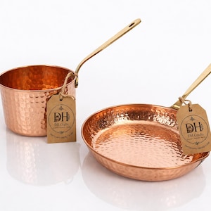 May include: A hammered copper saucepan and a matching frying pan, both featuring long brass-toned handles. Each piece has a brown paper tag attached with twine, printed with the text DH Crafts, Crafted with Heritage & Care.