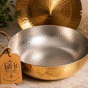 May include: A round, hammered metal serving bowl with a silver-toned interior and a gold-toned exterior. It features two gold-toned curved handles on the sides and a brown paper tag attached with string, printed with the text DH Crafts and DH.