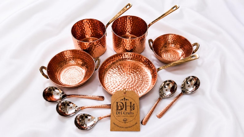 May include: A set of hammered copper cookware including two saucepans with long handles, two bowls with side handles, one frying pan, and four spoons with metallic bowls. A brown rectangular tag features the text DH Crafts and DH logo.