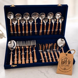 May include: A full set of copper and silver-coloured cutlery presented in a blue velvet case. The set includes spoons, forks, and knives. The handles have a hammered texture. A tag with the text "DH Crafts" is attached.