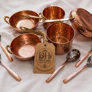 May include: A set of hammered copper cookware including two small bowls with side handles, two pots with long handles, and a lid, accompanied by four silver-toned spoons with hammered copper handles and a brown tag labeled DH Crafts.