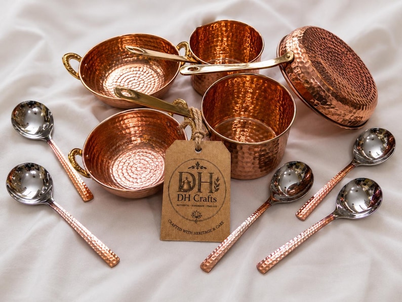 May include: A set of hammered copper cookware including two small bowls with side handles, two pots with long handles, and a lid, accompanied by four silver-toned spoons with hammered copper handles and a brown tag labeled DH Crafts.