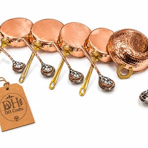 May include: A set of four small copper pans with brass handles, each paired with a matching metal spoon. A larger hammered copper bowl with a brass handle and a separate spoon are included. A brown paper tag features the text DH Crafts.