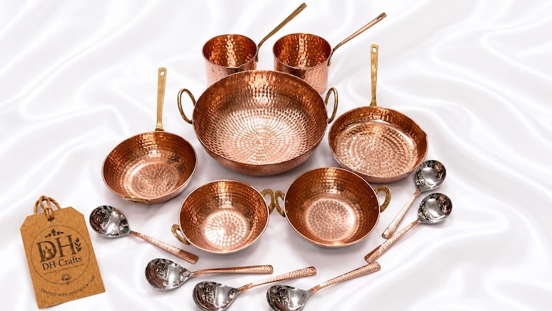 May include: A set of hammered copper cookware including a large central bowl, two smaller bowls with side handles, two saucepans with long handles, and six silver-toned spoons. A brown cardboard tag with the text DH Crafts is included.