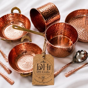 May include: A collection of hammered copper kitchenware including two bowls with brass handles, a saucepan with a long brass handle, a cylindrical cup, a frying pan, and four spoons with silver-toned bowls and hammered copper handles. A tag reads DH Crafts.