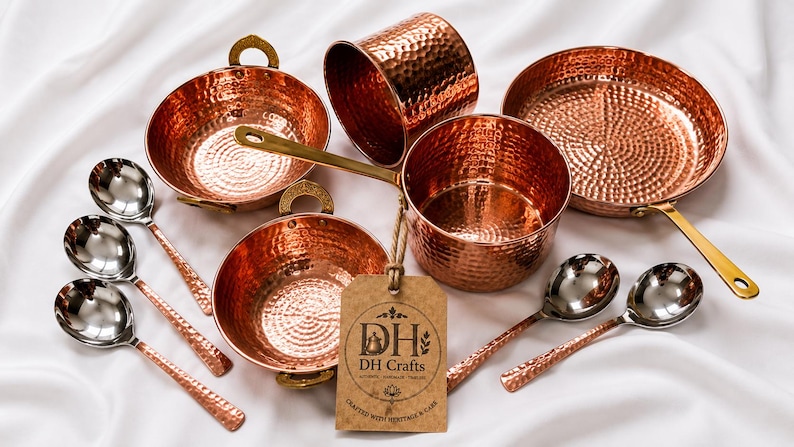 May include: A collection of hammered copper kitchenware including two bowls with brass handles, a saucepan with a long brass handle, a cylindrical cup, a frying pan, and four spoons with silver-toned bowls and hammered copper handles. A tag reads DH Crafts.