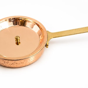 May include: A round copper pan with a hammered exterior finish and a smooth interior surface. The pan features a long, flat brass handle attached with rivets and a small, centred brass knob on the lid. The metal surfaces are polished and reflective.