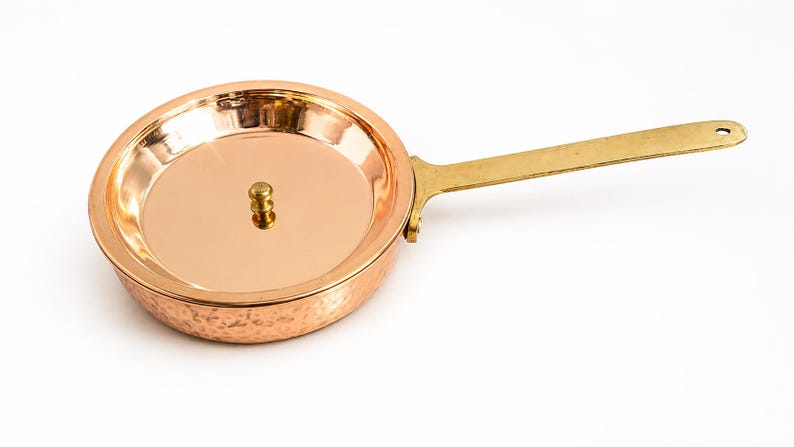May include: A round copper pan with a hammered exterior finish and a smooth interior surface. The pan features a long, flat brass handle attached with rivets and a small, centred brass knob on the lid. The metal surfaces are polished and reflective.
