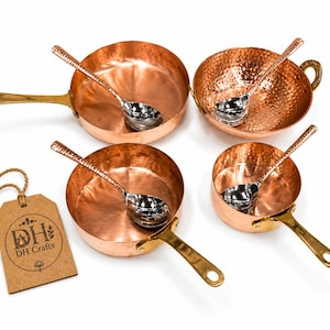 May include: A set of four copper pans and bowls with hammered textures, each paired with a silver-toned spoon. The pans feature brass handles. A brown cardboard tag with a jute string is attached, displaying the text DH Crafts in a circular logo.