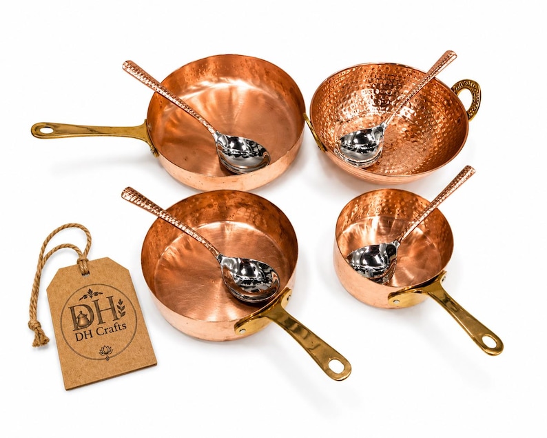 May include: A set of four hammered copper pans and bowls with brass handles, each paired with a silver-toned metal spoon. A brown cardboard tag with the text DH Crafts is attached by a jute string to the side of the collection of cookware.