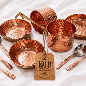 May include: A set of hammered copper kitchenware including two bowls, two pots, and five spoons with metallic handles. A brown cardboard tag is attached to one pot with twine, featuring the printed text DH Crafts and Crafted with Heritage & Care.