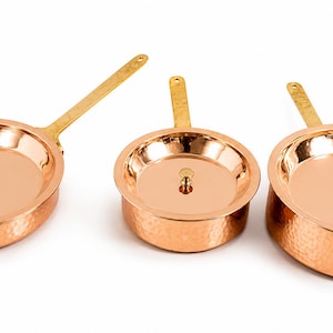 May include: A set of three round, hammered copper pans with polished brass handles and matching lids. Each lid features a small, centred brass knob. The exterior surfaces of the pans exhibit a textured, dimpled finish, while the interior surfaces are smooth and reflective, showcasing the metallic copper tone.