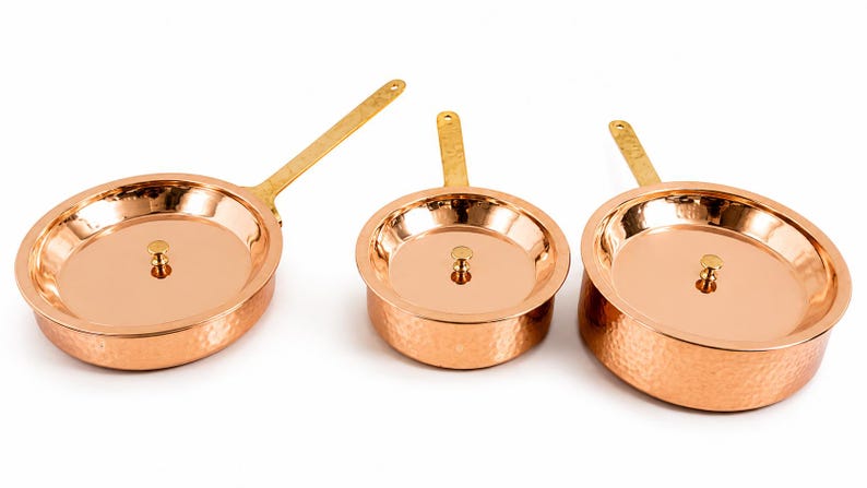 May include: A set of three round, hammered copper pans with polished brass handles and matching lids. Each lid features a small, centred brass knob. The exterior surfaces of the pans exhibit a textured, dimpled finish, while the interior surfaces are smooth and reflective, showcasing the metallic copper tone.