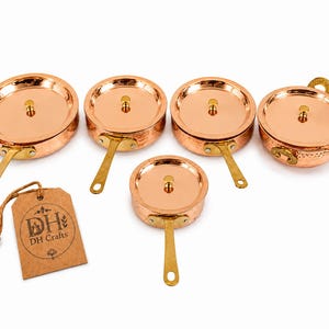 May include: A set of four round copper pans and one bowl with lids, each featuring a brass handle and a hammered texture. A brown paper tag is attached by a string, displaying the text DH Crafts with a small graphic of a pot and floral accents.