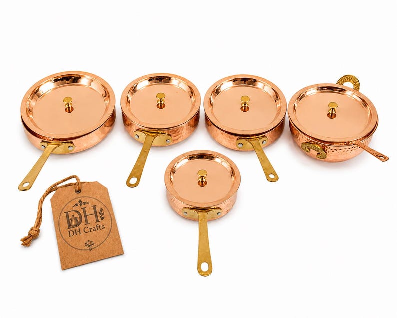 May include: A set of four round copper pans and one bowl with lids, each featuring a brass handle and a hammered texture. A brown paper tag is attached by a string, displaying the text DH Crafts with a small graphic of a pot and floral accents.