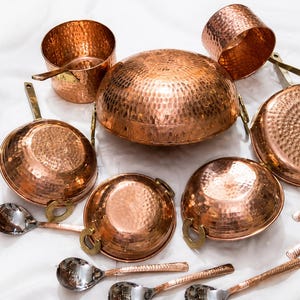 May include: A set of hammered copper cookware including a large central dome-shaped pot, several smaller bowls with brass handles, and silver-toned spoons. A brown cardboard tag with the text DH Crafts and Crafted with Heritage & Care is included.