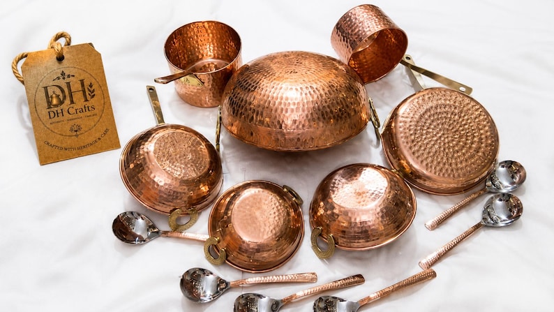 May include: A set of hammered copper cookware including a large central dome-shaped pot, several smaller bowls with brass handles, and silver-toned spoons. A brown cardboard tag with the text DH Crafts and Crafted with Heritage & Care is included.