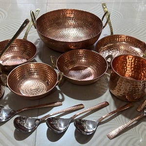 May include: A collection of hammered copper cookware including a large round bowl with two handles, two smaller bowls with handles, two deep pots, and a shallow pan. Five silver-toned metal spoons with textured handles are arranged in the foreground.