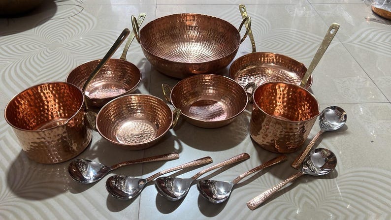 May include: A collection of hammered copper cookware including a large round bowl with two handles, two smaller bowls with handles, two deep pots, and a shallow pan. Five silver-toned metal spoons with textured handles are arranged in the foreground.