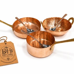 May include: A set of three copper cookware pieces including two pans with long brass handles and one bowl with two brass side handles. Each piece contains a silver-toned spoon. A brown paper tag features the text DH Crafts, Crafted with Heritage & Care.