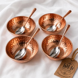 May include: Four round, hammered copper bowls, each paired with a polished silver-tone metal spoon. A brown paper tag with the printed text DH Crafts and a lotus flower icon is attached to the set by a thin, light brown twine string.