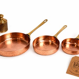 Op de afbeelding: Drie ronde koperen pannen met een gehamerde textuur en messing handgrepen, gerangschikt in afnemende grootte. Aan de kleinste pan hangt een bruin papieren label met de tekst DH Crafts, Crafted with Heritage & Care.