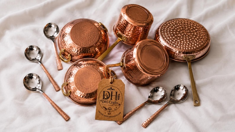 May include: A collection of hammered copper cookware including two round pots with side handles, two saucepans with long handles, and five silver-toned spoons with textured copper handles. A brown paper tag displays the text DH DH Crafts.
