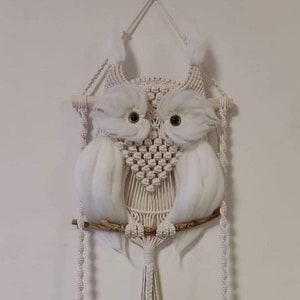 Könnte beinhalten: Ein weißer Makramee-Eulen-Wandbehang mit einem Holzstab und weißen Garnquasten. Die Eule hängt über einem Holzregal mit einem schwarzen Keramiktopf, einer goldenen Vase und einer Muschel.