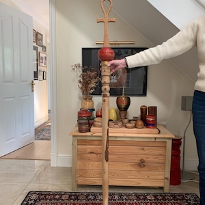 Può includere: Un bastone Ankh in legno alto con una parte superiore a forma di croce e una sfera rossa. Il bastone è marrone chiaro con un design intagliato e una serie di perline di legno. Il bastone è alto circa 152 cm.