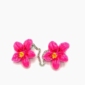 May include: A pair of pink flower-shaped brooches connected by a silver chain. The flowers have yellow centers and red speckles.