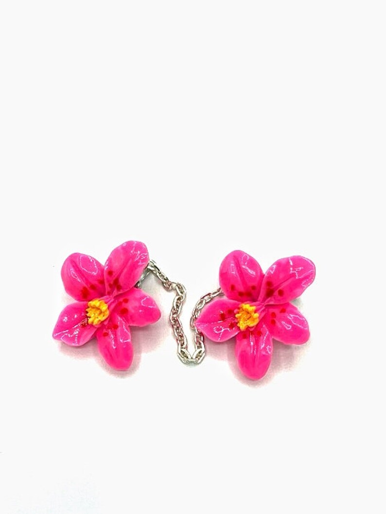 May include: A pair of pink flower-shaped brooches connected by a silver chain. The flowers have yellow centers and red speckles.