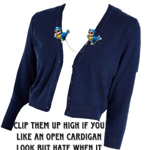 May include: A navy blue cardigan with two blue bird clips pinned to the neckline. The text "CLIP THEM UP HIGH IF YOU LIKE AN OPEN CARDIGAN LOOK BUT HATE WHEN IT BLOWS IN THE WIND." is below the image.