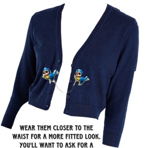 May include: A navy blue cardigan with two blue bird clips attached to the front. The text "EXAMPLE OF HOW TO WEAR CLIPS." is at the top of the image. The text "WEAR THEM CLOSER TO THE WAIST FOR A MORE FITTED LOOK. YOU'LL WANT TO ASK FOR A SHORTER CHAIN." is at the bottom of the image.
