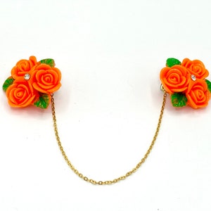 May include: A pair of orange rose brooches connected by a gold chain. Each brooch features three orange roses with green leaves and a single rhinestone in the center.