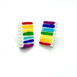 May include: A pair of rainbow cake slice earrings. The earrings are made of white polymer clay and feature a rainbow design with red, orange, yellow, green, blue, indigo, and violet stripes.