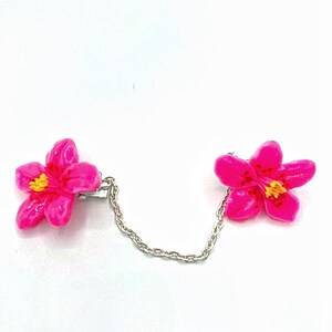 May include: A pair of pink flower shaped clips connected by a silver chain. The flowers have yellow centers.