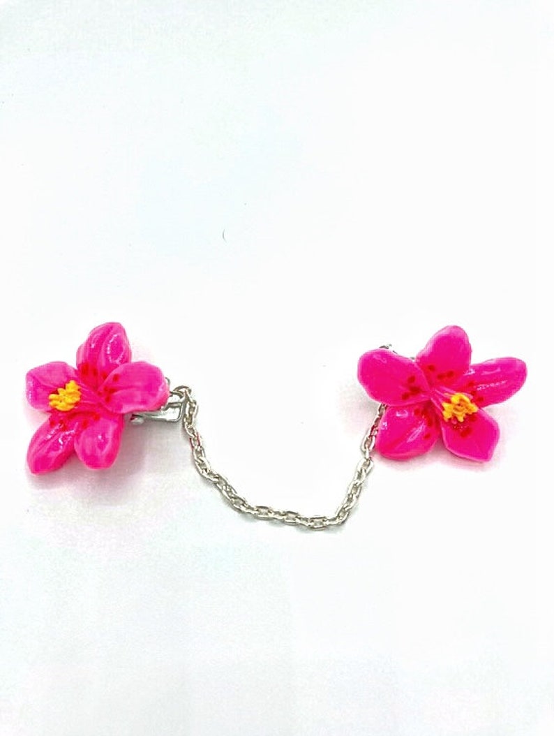 May include: A pair of pink flower shaped clips connected by a silver chain. The flowers have yellow centers.