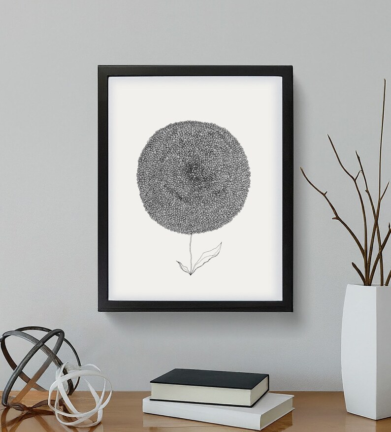 May include: Black framed art print featuring a detailed, grayscale illustration of a dandelion-like flower. The artwork is mounted on a white background. The image is displayed on a wooden surface with decorative objects.