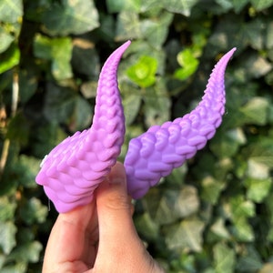 May include: Two lavender-colored, textured, horn-shaped objects are held in front of a green leafy background. The horns have a curved shape and a detailed, scale-like texture, with a lighter shade of lavender at the tips.