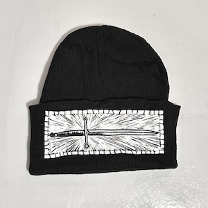 May include: Black beanie hat with a white rectangular patch featuring a sword design. The sword is centered, with rays emanating from it. The design is stitched onto the hat.