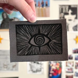 May include: A gray fabric patch featuring a black eye design with radiating lines. The eye has a spiral pupil. The patch is rectangular with a gray border. The design is a linocut style.