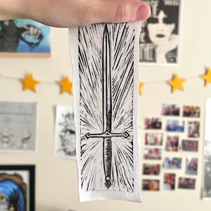 May include: A white rectangular fabric with a black sword design. The sword has a long blade and a cross-shaped hilt. The design is a linocut print. The fabric is held up in front of a wall with artwork and star-shaped lights.