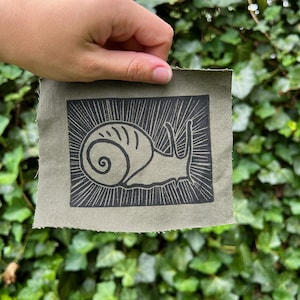 May include: Olive green fabric patch featuring a black linocut print of a snail with radiating lines. The snail design is centered on the patch, creating a simple, yet striking image. The patch has a raw, unfinished edge.