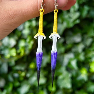 May include: A pair of yellow, white, purple, and black dagger earrings. The earrings are made of plastic and have a silver hook.