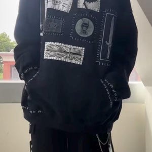 May include: A black hooded sweatshirt featuring a series of white rectangular patches on the front. The patches display various graphic illustrations including a skull, a bat, an eye, a sword, and a circular emblem. White text is printed on the sleeves.