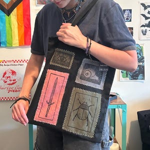 May include: A black tote bag featuring four rectangular patches with hand-stitched edges. The patches display a central eye, a snail, a sword on a pink background, and a beetle. The bag includes a small black tag with the text S M L XL XXL.