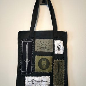 Puede incluir: Bolso tote negro con varios parches con diseños de espada, calavera, araña, perro, cruz y hoja curva. Los parches están cosidos a la bolsa. La bolsa tiene asas negras.