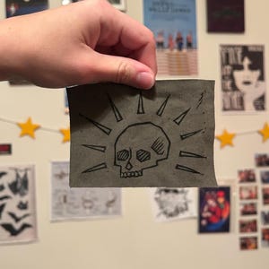 May include: A gray fabric patch featuring a black skull design with radiating lines. The skull has detailed eye sockets and teeth. The patch is held up against a wall with various posters and decorations.
