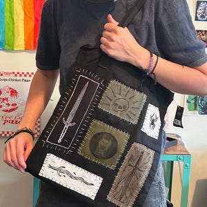 May include: A black tote bag featuring several rectangular fabric patches sewn onto the front. The patches display white and black illustrations of a long sword, a skull with radiating lines, a spider, a snarling animal head, and a wavy blade.